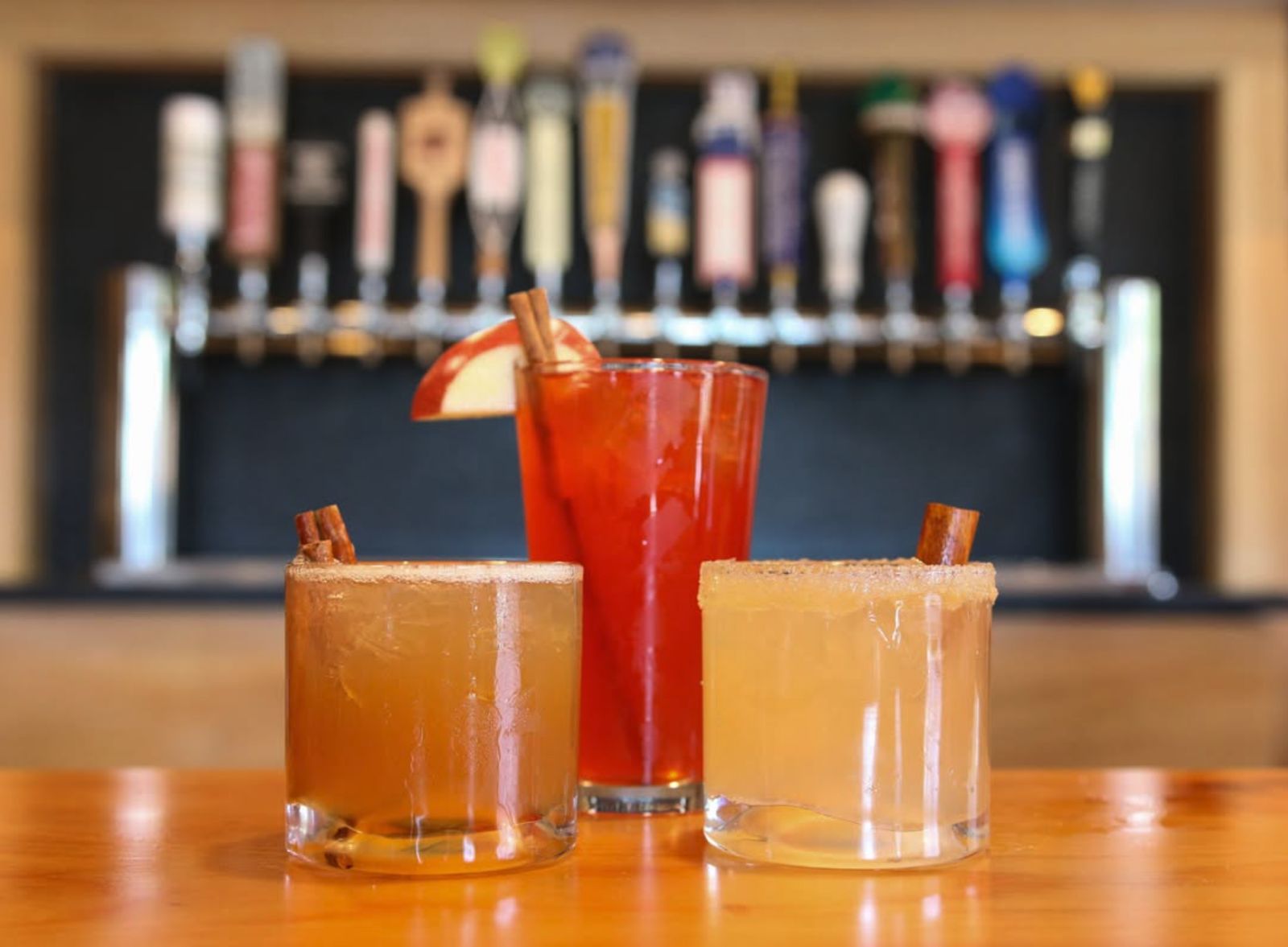 Three fall cocktails with cinnamon sticks in front of Alpine Pizza's tap wall