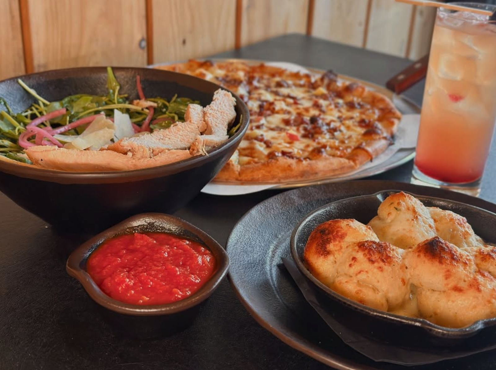 A pizza, arugula salad with chicken, garlic knots, and a cocktail on a table